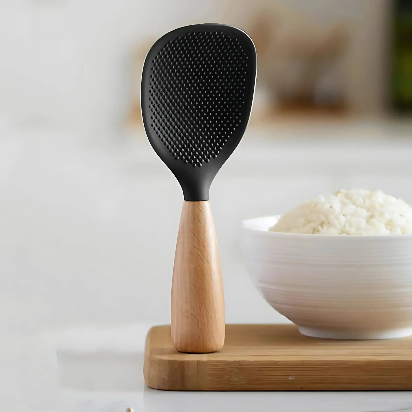 Black rice paddle with wooden handle on a white surface with a bowl of rice in the background