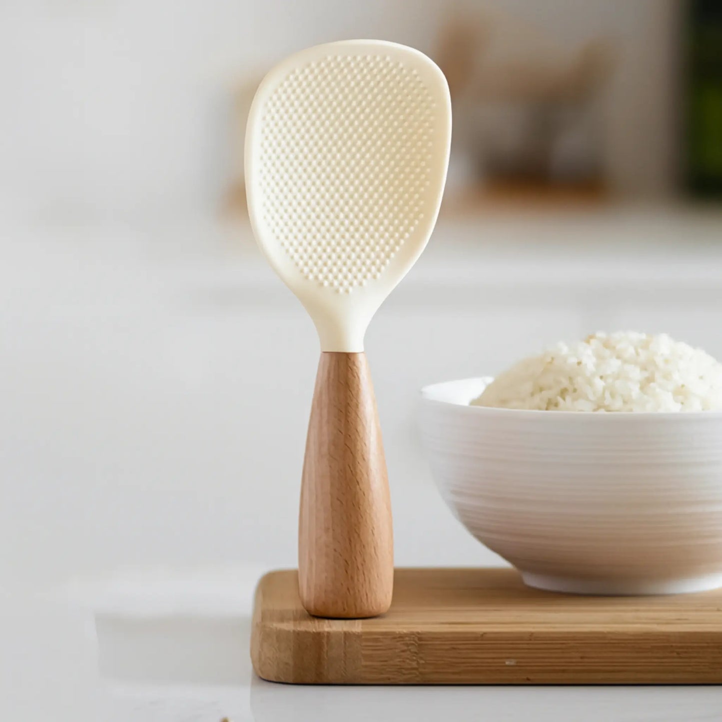 White rice paddle with wooden handle on a wooden block, next to a bowl of rice.