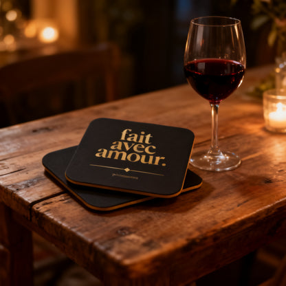 Two Fait Avec Amour black and gold coasters stacked on rustic wooden table with glass of red wine and candlelight - petitegourmets